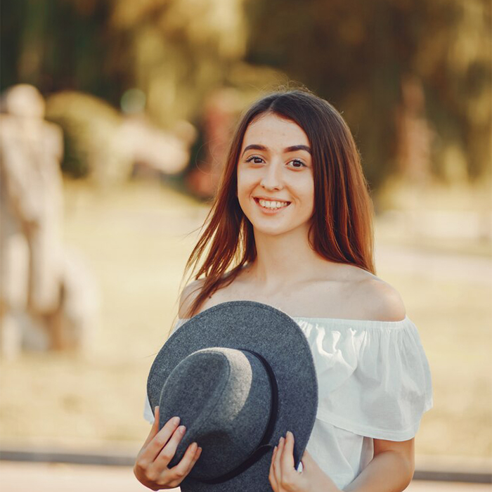 Charlotte smiling outdoors while holding a gray hat, wearing an off-the-shoulder white top.